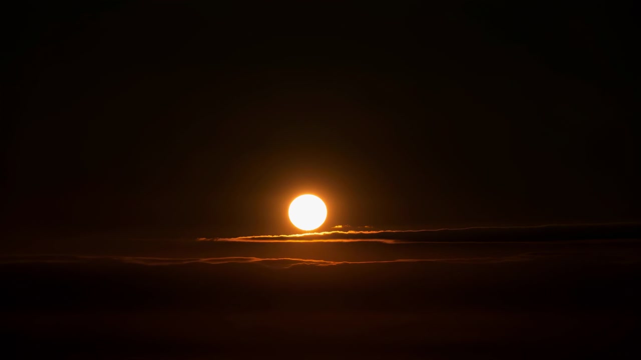 Lave skybånd forbliver silhuetteret under en ekspanderende ravfarvet glød, mens den cirkulære sol stiger fra horisonten op på en åben mørk himmel, hvilket skaber stærk kontrast og en centreret atmosfærisk udsigt