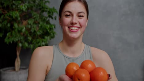 Portrait-of-a-confident-and-happy-young-brunette-girl-in-a-gray-sports-top-who-holds-four-orange-oranges-in-her-hands-near-a-large-indoor-plant-in-a-modern-apartment-at-home