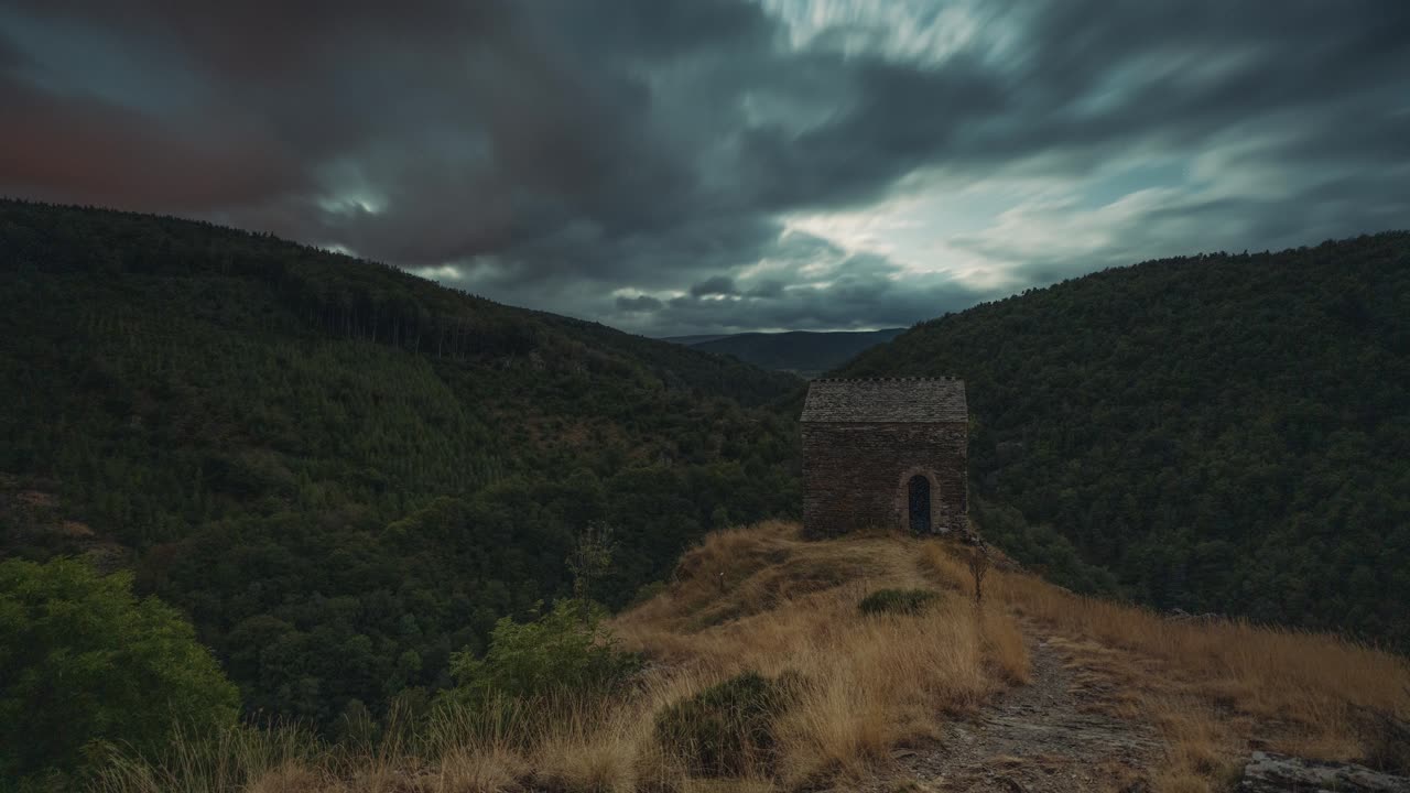Night timelapse of a moonlit, ancient stone house on a hilltop overlooking a dense, green valley. Soft clouds diffuse the moonlight, casting a mystical glow over the landscape.
