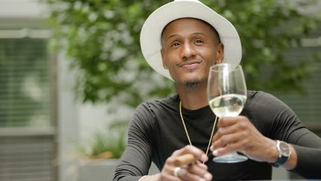 happy wealthy man wearing fedora hat and gold jewelry sits at a table swirling and drinking wine outside near green plants