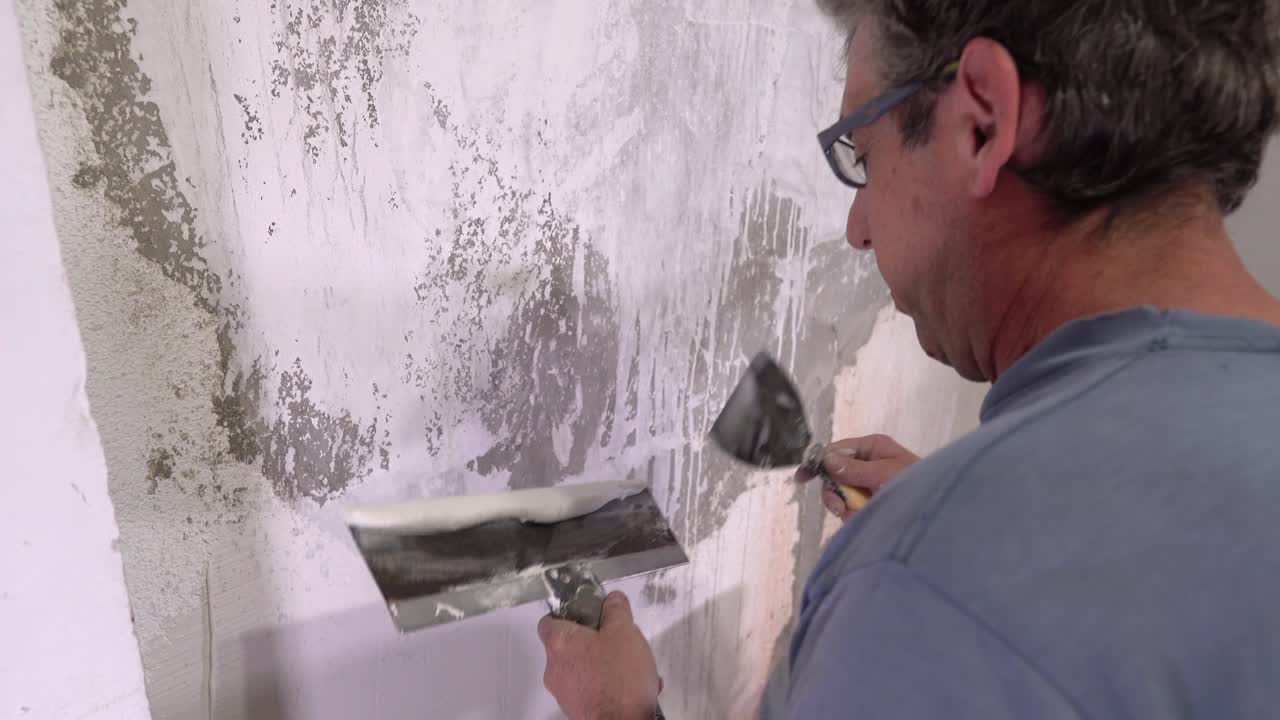 Manual wall finishing technique is demonstrated as a worker uses a large metal taping knife to smooth joint compound indoors