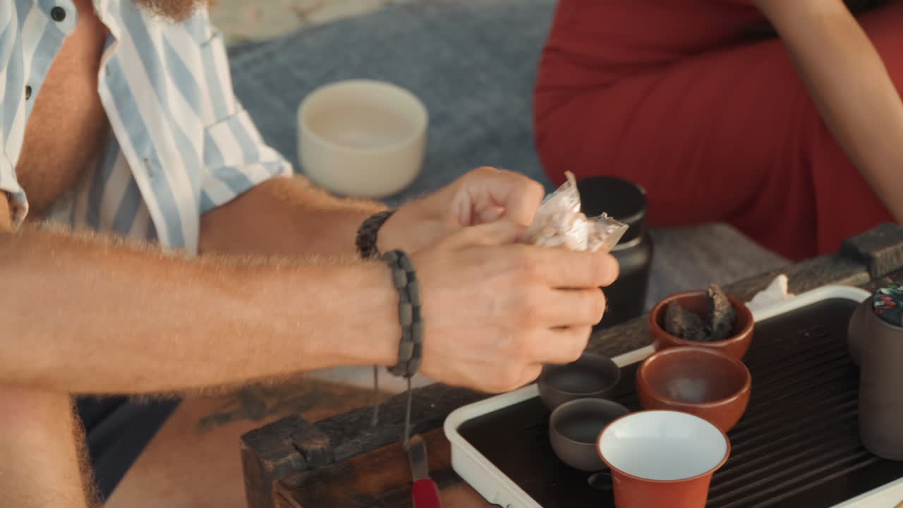 Pareja en ceremonia del té en la playa preparando hojas sueltas en una bandeja, hombre manipulando tazas con delicadeza, pareja sentada en una manta con vestido rojo, teteras de cerámica y hervidor en bandeja de madera, vapor ascendiendo, luz solar dorada, océano