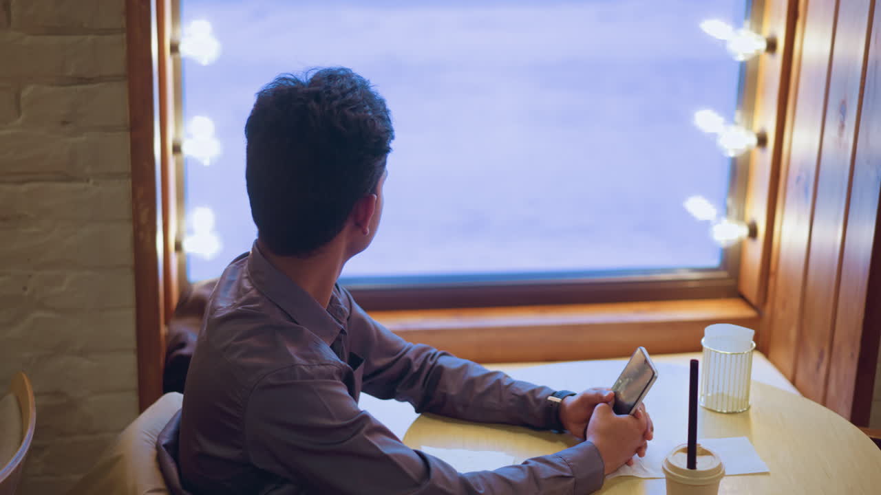 Man in casual shirt sitting near cafe window holding phone, staring outside thoughtfully with coffee cup on table, warm indoor light contrasting cold snowy outdoor scene, evoking solitude and contemplation