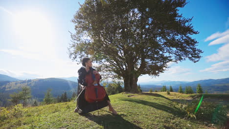 female music player sitting on the chair plays violoncello. classical music player at backdrop of mountains on sunny day.