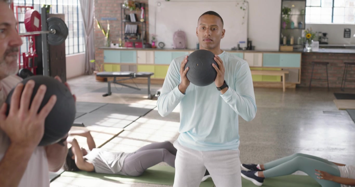 Diverse group following center man, holding black medicine balls and squatting for fitness in loft
