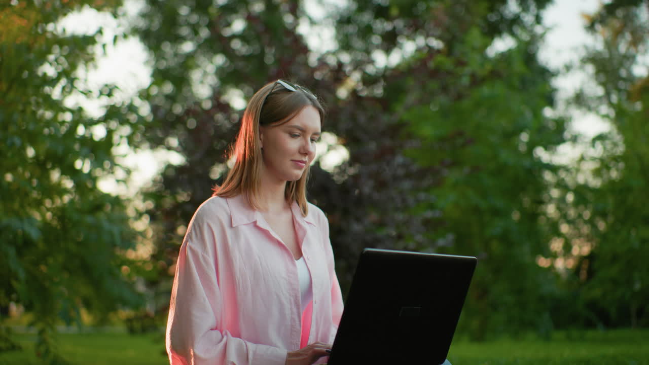 joven adulto en rosa sentado con las piernas cruzadas en un campo de hierba, trabajando en su portátil, mira hacia arriba pensativo con una sonrisa cálida, el fondo presenta árboles y vegetación
