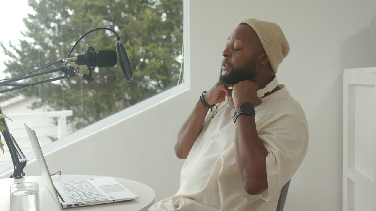 Adult African man reaching adjusting mic pop-filter at laptop donning headphones prepping to record
