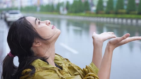 젊은 아름다운 아시아 여성은 노란색 레인코트의 후디를 입고 손을 사용한 후 떨어지는 비를 잡고, 비가 오는 계절의 날씨, 폭풍우의 슬, 불행한 날, 불행, 뒷면의 도시 거리