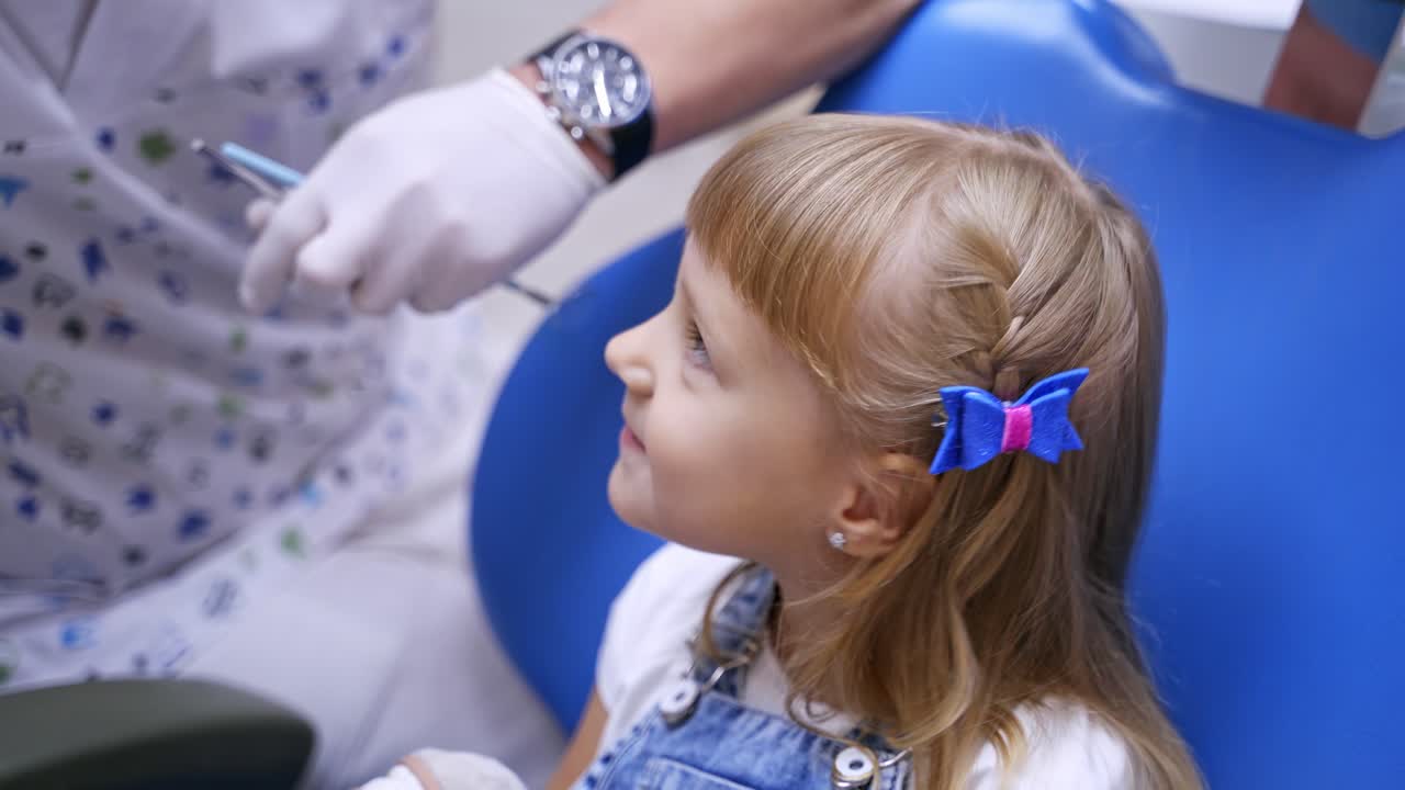 Little girl sitting in dentists chair. Pretty kid opens her mouth to the stomatologist. Doctor checks girl's teeth.