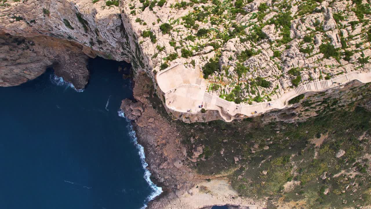 A cliff with a path leading to a viewpoint. Tourists enjoy the view at Cap de Formentor