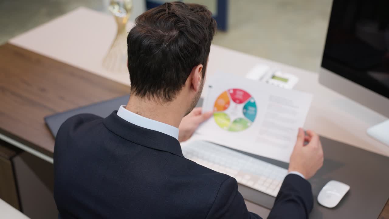 A businessman CEO is reviewing a colorful chart document while seated at his desk in a modern office environment, focusing on strategic planning or analysis - over shoulder view parallax shot
