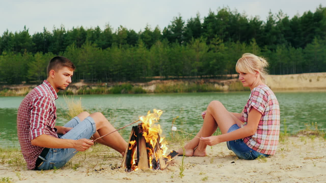 한 남자와 한 여자의 친구가 모닥불 주위에 앉아 막대기에 마시멜로를 굽고 있습니다.