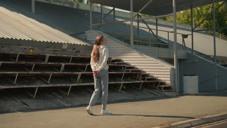 lady in sporty attire, and long hair tied back walks thoughtfully near a stadium on a sunny day, with hands behind her back, she appears deep in thought, surrounded by empty stadium seating