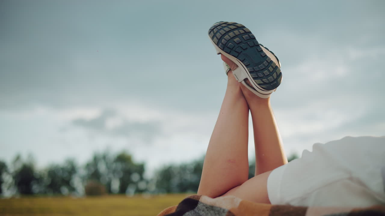 에 누워있는 쇼트를 입은 여성의 측면 뷰, 다리를 들어 올리고 샌달을 고, 구름이 은 하늘과 흐릿한 배경은 평화로운 야외 분위기를 만니다.