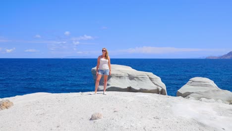 pretty woman on the edge of the sarakiniko cliffs with ocean in the background turns to face the camera in slow motion