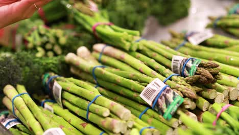 a hand picks asparagus bundles in a well-lit supermarket produce section, highlighting fresh greens and careful selection