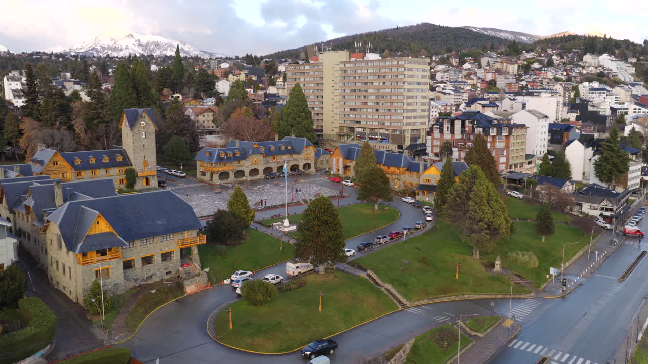 Descending daisy cutter drone shot approaches city plaza and buildings with lake and snowy mountains behind