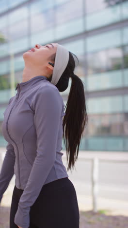 vertical video of young woman exercising running along city street wearing wireless earbuds resting