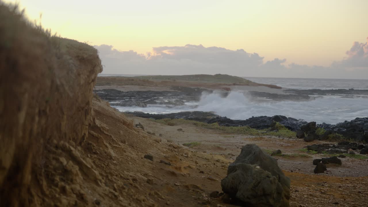 A blurred man in white holds a smartphone off-center in the frame, as the rocky coastline and ocean horizon dominate under the glow of sunrise