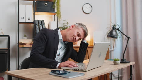 exhausted young businessman in formal suit working on laptop computer sitting at desk in home office