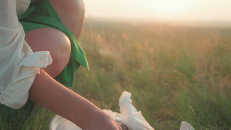 close up of woman kneeling on grass while unpacking food and supplies from canvas tote bag for picnic, with plastic container, fruit, and wildflowers visible in soft natural light at sunset