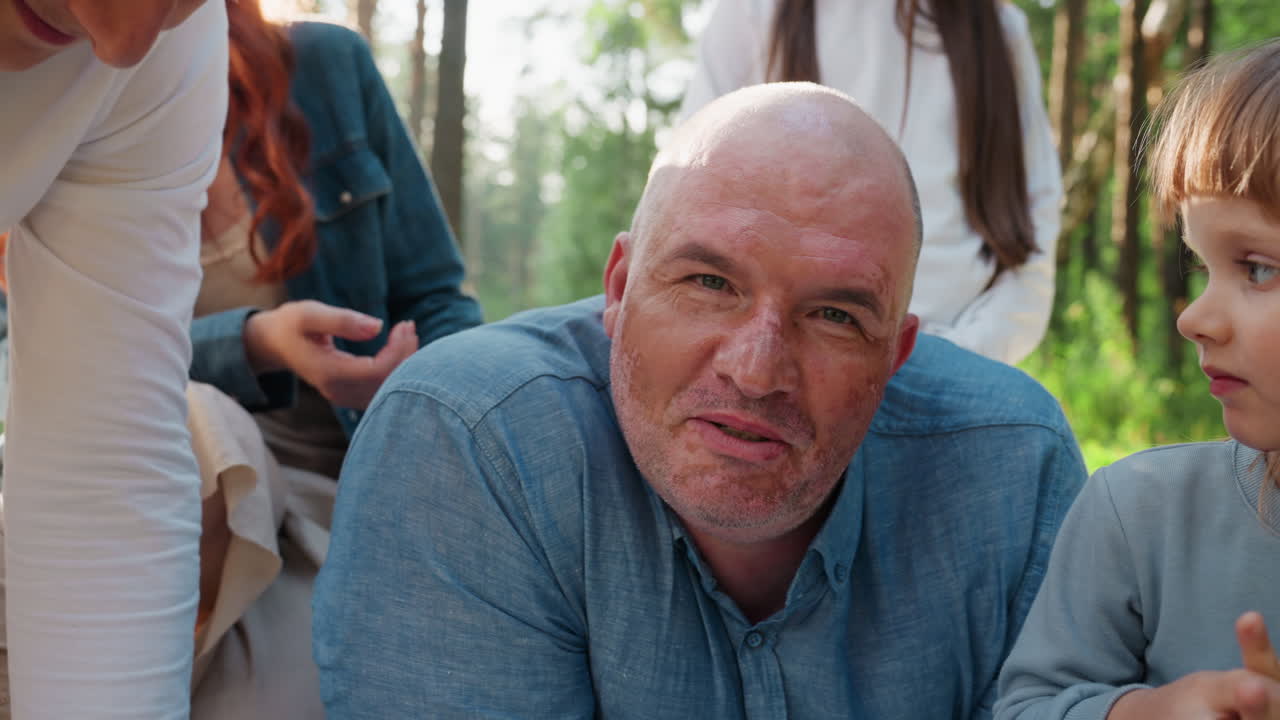 Close up of dad lying comfortably on mat surrounded by loving family in warm sunlight, sharing peaceful outdoor moment together in forest setting, showing tenderness, and relaxation