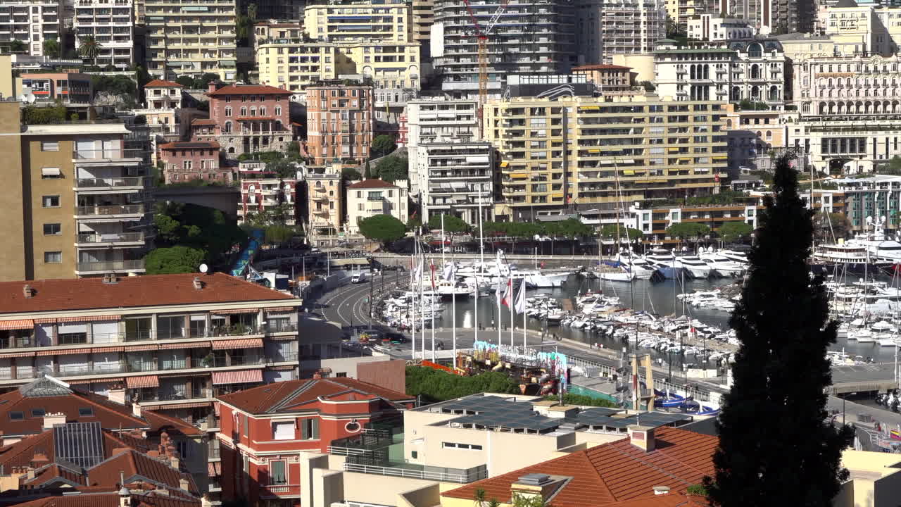 La Condamine, Monaco - July 4, 2025: Busy curve of Quai Albert-1er with traffic, Monaco flags, a seasonal funfair, and rows of yachts moored in Port Hercule below residential blocks