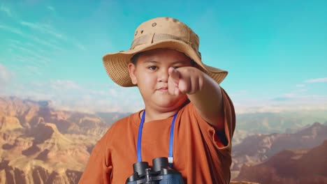 asian boy with hat and binoculars using magnifying glass pointing finger towards you, shaking head waving index finger while traveling at the top of mountain. boy researcher showing no gesture, close up