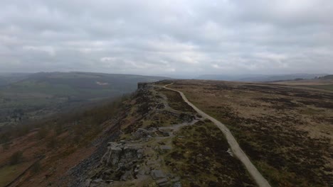 Aerial-drone-footage-flying-over-a-long-rural-trail-between-open-moorland-fields-and-dramatic-rock-formation