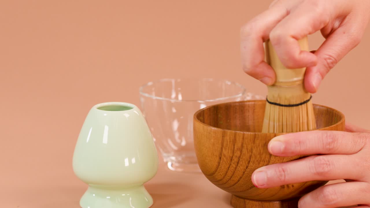 Hands whisk matcha in wooden bowl using bamboo chasen, soft lighting, neutral background, close-up