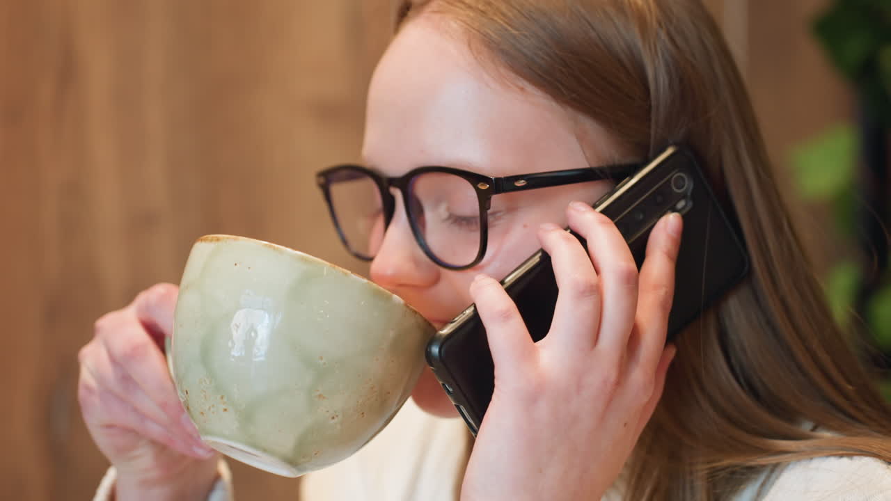 close up cheerful lady wearing glasses chatting on phone with friend while enjoying coffee moment in cozy indoor setting smiling warmly expressing joy during relaxed personal conversation