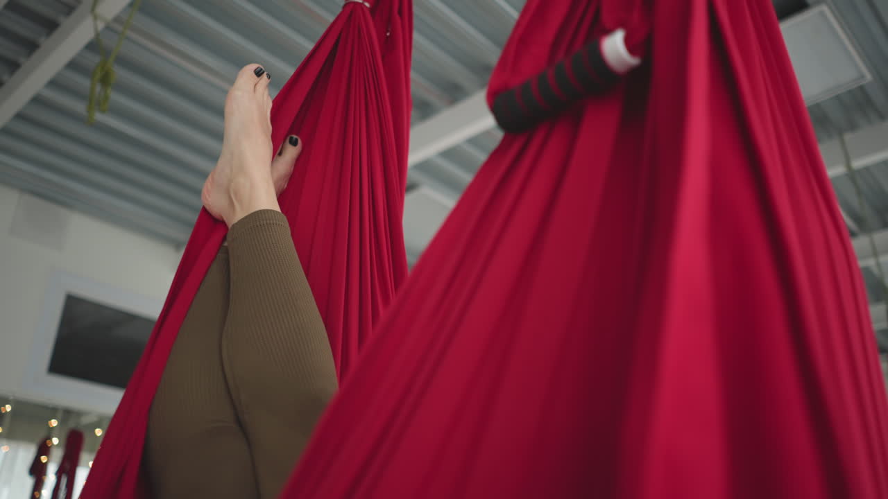 Flexible woman resting in red silk hammock swinging gently above floor mat with feet visible and serene smile bright studio with sheer curtains and urban window view conveying balance and calm
