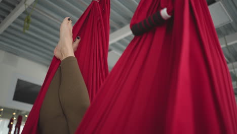 flexible woman resting in red silk hammock swinging gently above floor mat with feet visible and serene smile bright studio with sheer curtains and urban window view conveying balance and calm