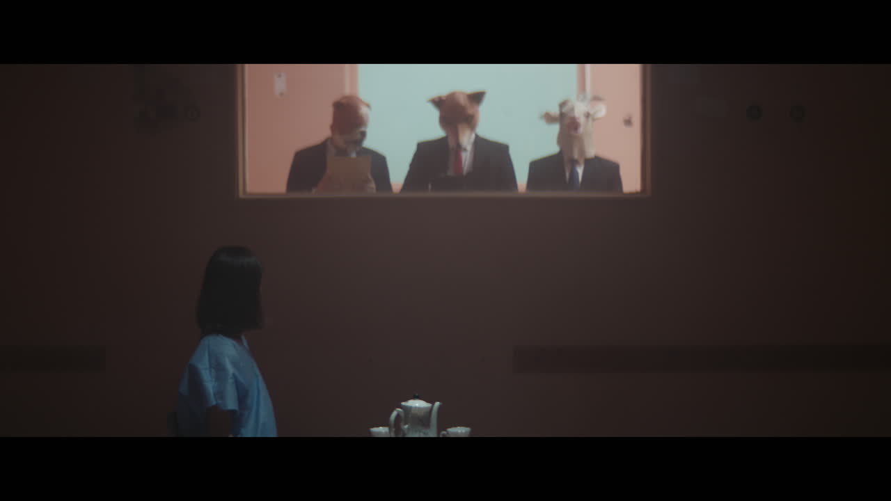 A girl sits at a table and strange figures in animal masks observes her from behind the glass