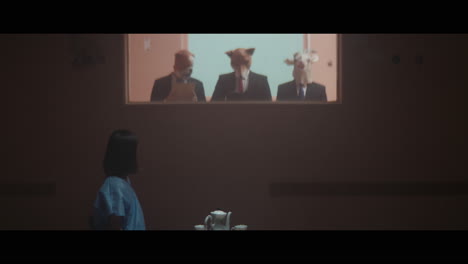 a girl sits at a table and strange figures in animal masks observes her from behind the glass