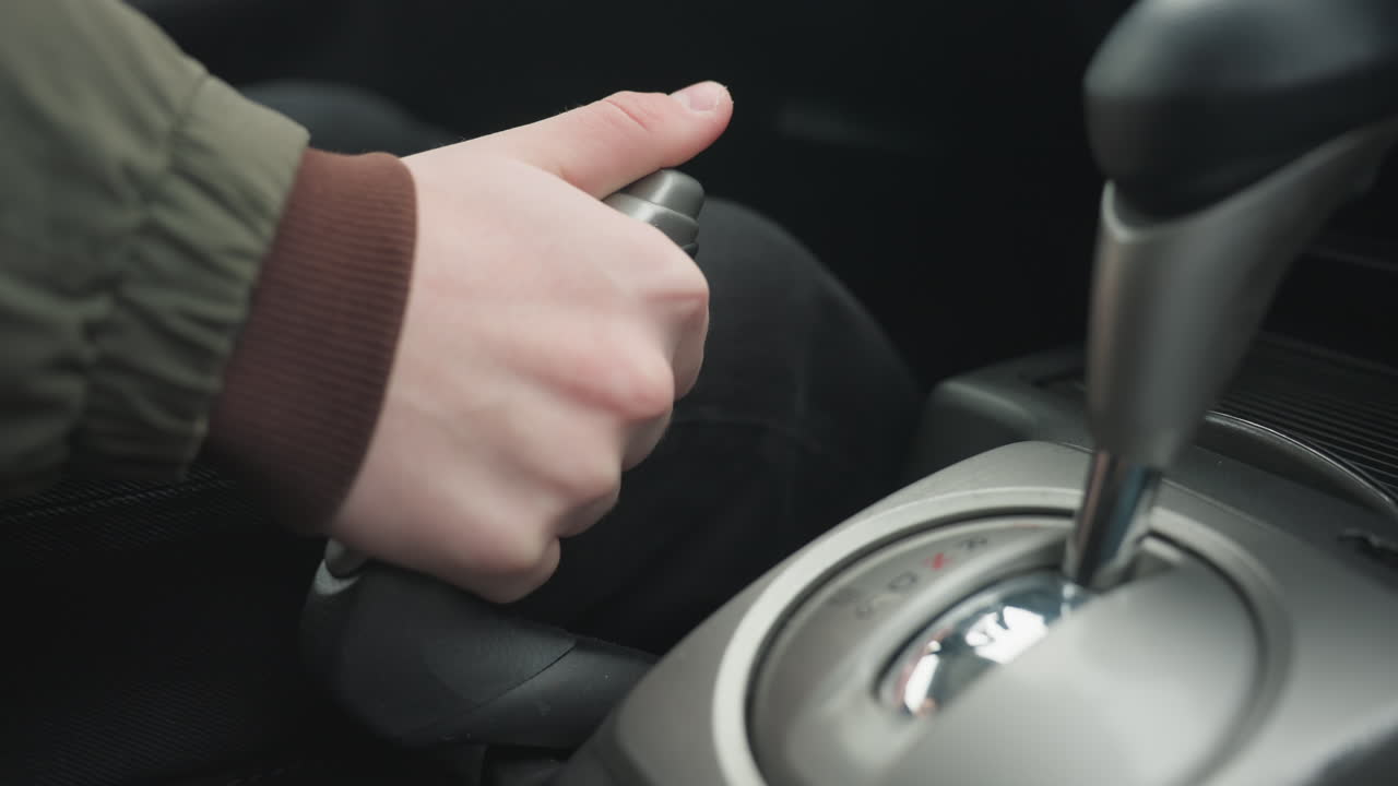 Close up of person pulling up hand brake lever on car center console showing hand gripping latch mechanism and leather trim under ambient daylight inside modern vehicle cabin detail