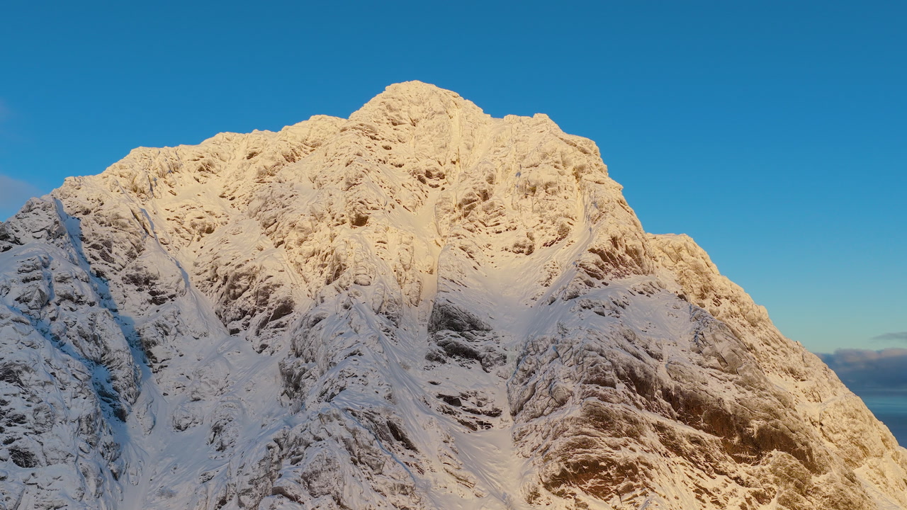 Dramatic drone shot of a rugged snowcapped mountain summit glowing in golden morning light against a clear blue sky in Lofoten, Norway.
