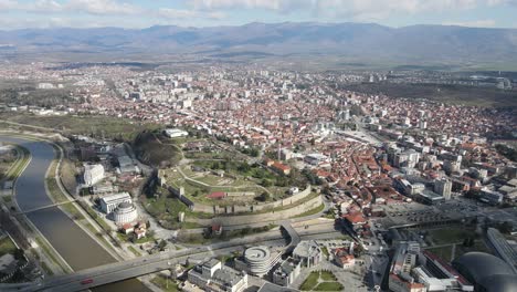 Vista-De-Drones-De-La-Ciudad-De-Skopje-Cruzando-El-Río-Vardar,-Castillo-De-Piedra-En-El-Centro-De-La-Ciudad