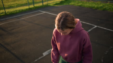 young lady in maroon sweater tossing volleyball in hand, bouncing ball with golden sunlight glow and green background, court markings visible, preparing for volleyball play or practice