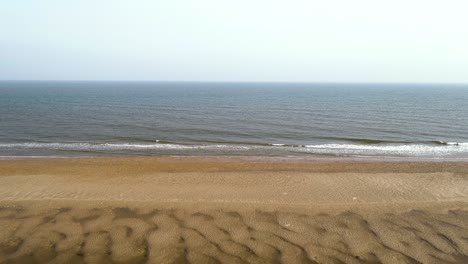 Ascending-drone-shot-of-beach-with-waves-and-sand-dunes-in-view-with-tide-out-and-open-water-looking-out-to-sea-in-Norfolk,-England