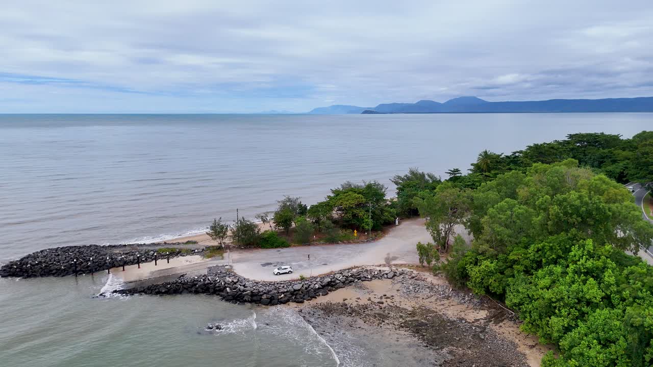 Drone glides above lush rainforest, rocky shoreline, and ocean under overcast daylight in Queensland