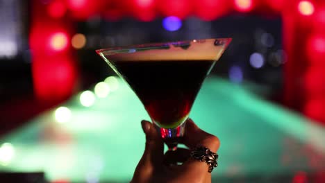 a hand rotates an espresso martini glass against a colorful, blurred nightclub background with dramatic red and green lighting and shallow depth of field