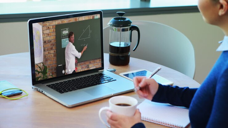 Estudiante caucásica usando una laptop en videollamada con un profesor, tomando apuntes.