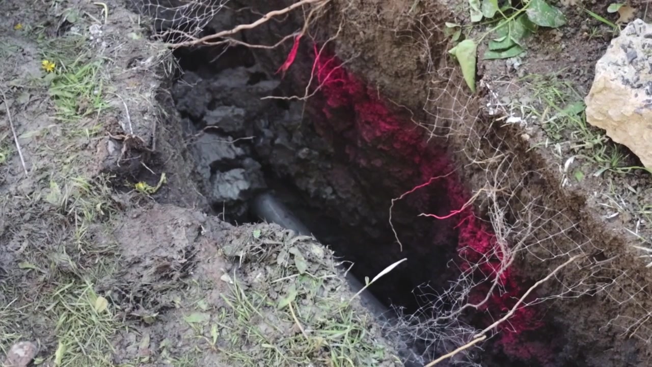 A slant‑face drill bit is pushed into an open hydrovac pit where soil, roots, mesh, and red electrical cables are exposed for street lighting conduit installation.