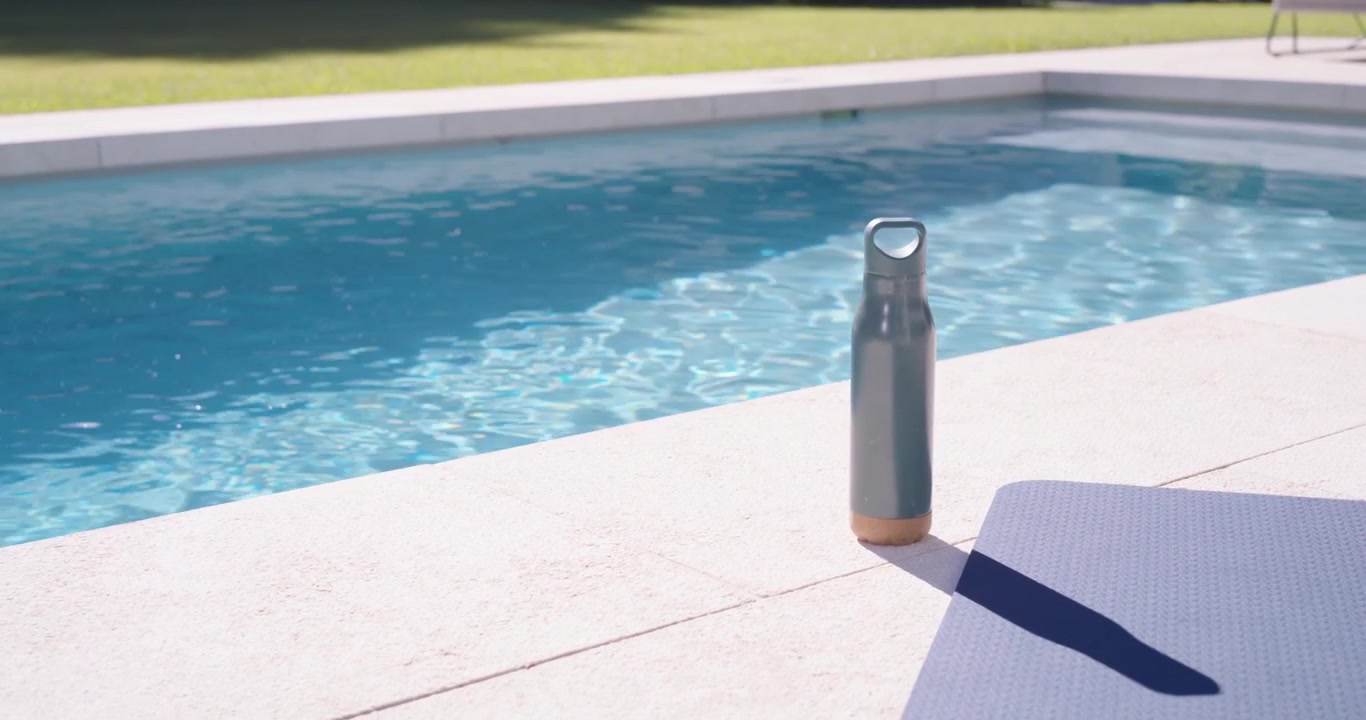 Metal water bottle is standing on pool deck while camera is pulling back, revealing sparkling pool