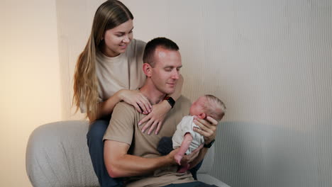 new-made dad looks at his newborn son with adoration holding him in hands. mom sits behind her husband.