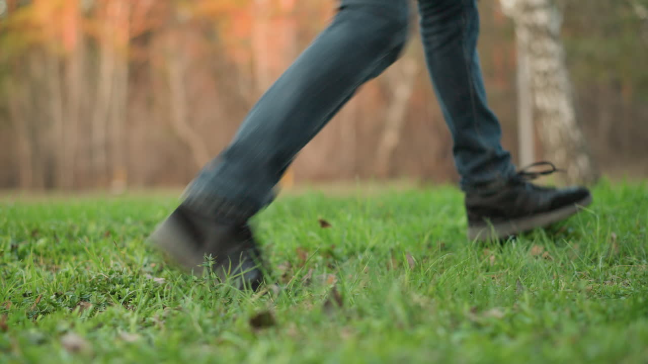 A close-up shot captures a man's legs in jeans and black shoes walking on lush green grass. The background shows a soft, blurred outdoor park setting with trees bathed in warm sunlight