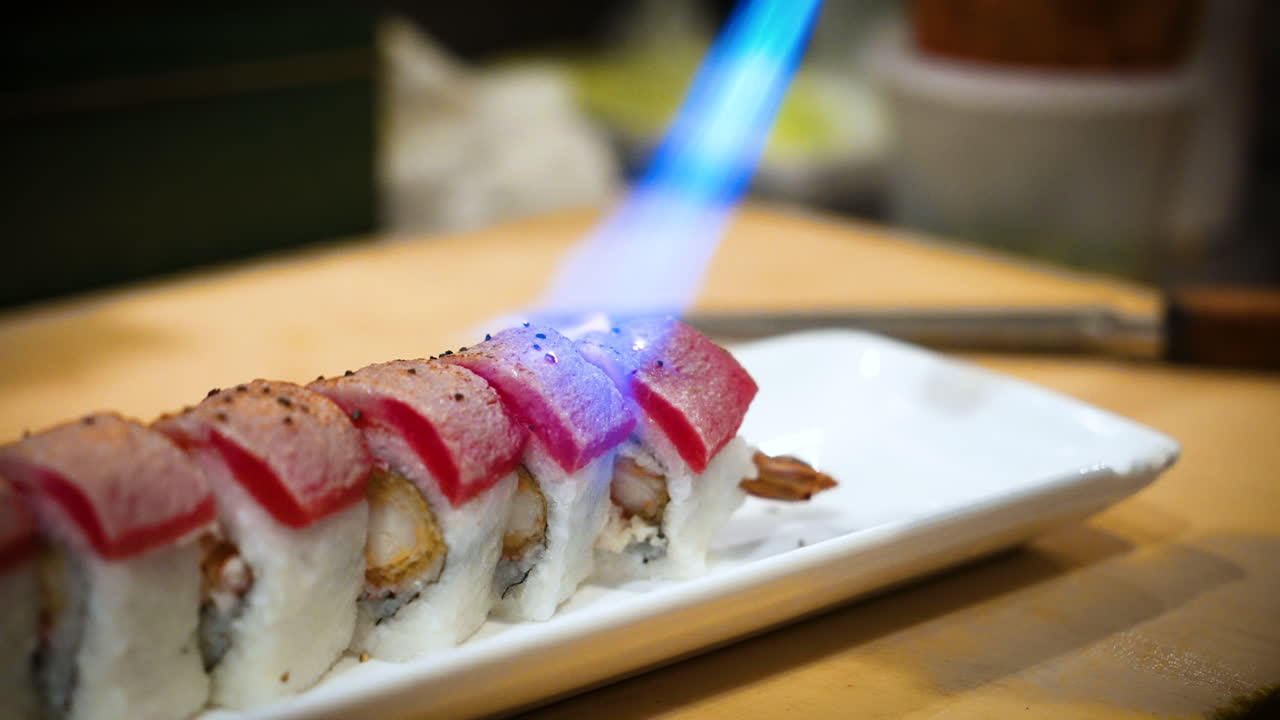 Chef uses hand torch to sear raw tuna on top of sushi roll, blue flame blasts fire cooking raw fish sushi, slow motion close up 4K