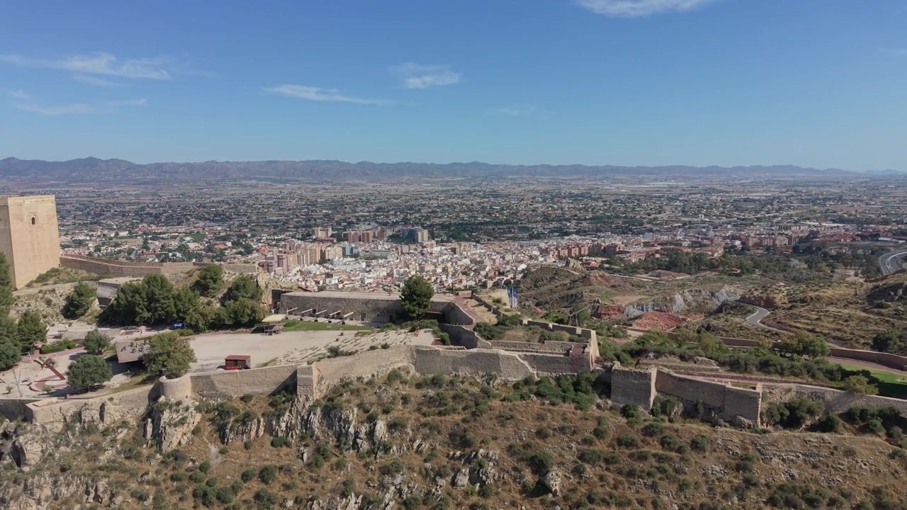 Vista aerea con drone delle fortificazioni del Castello di Lorca che dominano la storica città di Lorca, Spagna, con un'ampia espansione urbana, valli e paesaggi montani in lontananza.