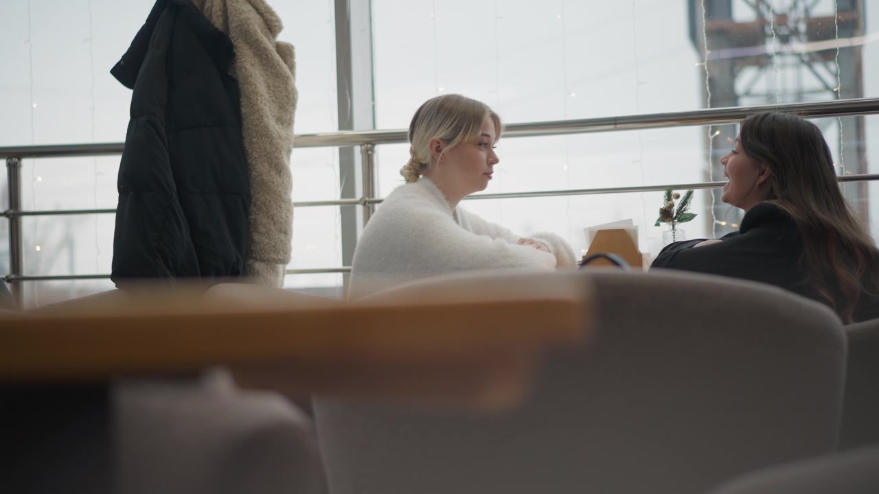 Two women sit facing each other across table in bright cafe, one leaning forward with hands near tabletop while both share calm attentive expressions and relaxed conversation, with women